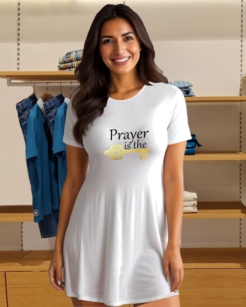 A woman standing in a clothes shop wearing a white t-shirt dress featuring a logo design by Heavenly Dreamwear.  The logo includes black text that reads, "Prayer is the," followed by a gold key symbol.