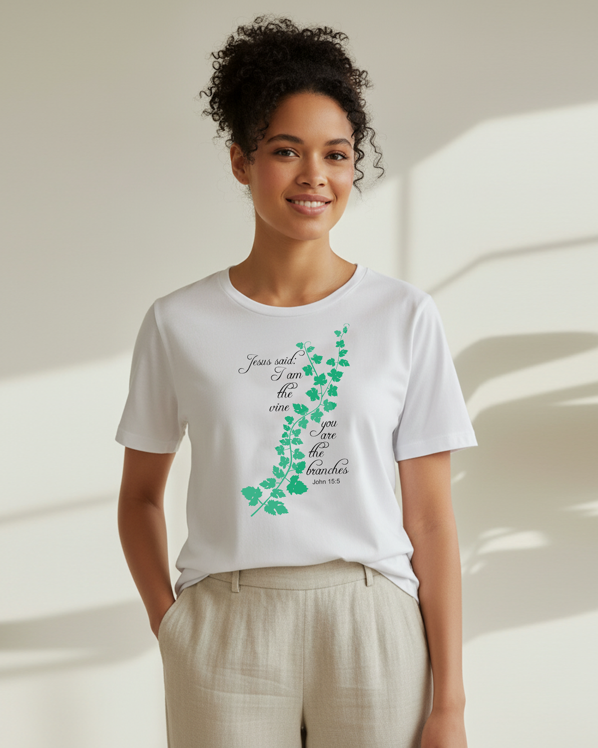 A woman wearing a white short-sleeve crewneck t-shirt with a green vine graphic and black cursive text, paired with light beige linen pants, standing in a brightly lit room with soft shadows.