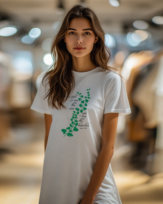 Woman standing in a shop wearing a white t-shirt dress with a logo made up of a green vine draped across the front with a biblical scripture on either side in black text from John 15:5