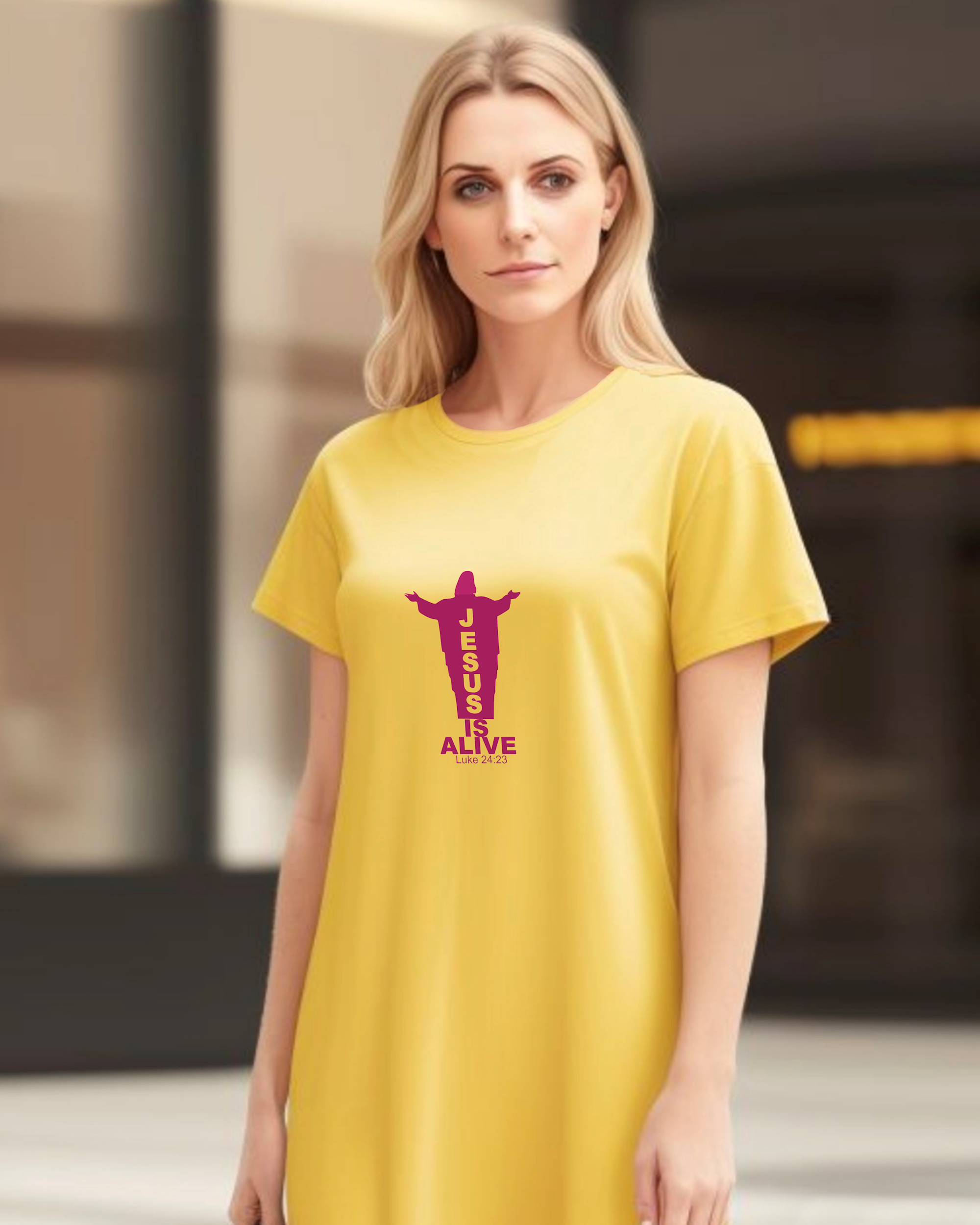 A woman in a garden wearing a yellow Heavenly Dreamwear t-shirt dress with a deep red logo of a silhouette of Jesus with the name "JESUS" embedded in it. Underneath is the text "IS ALIVE" with the scriptural reference: Luke 24:23.