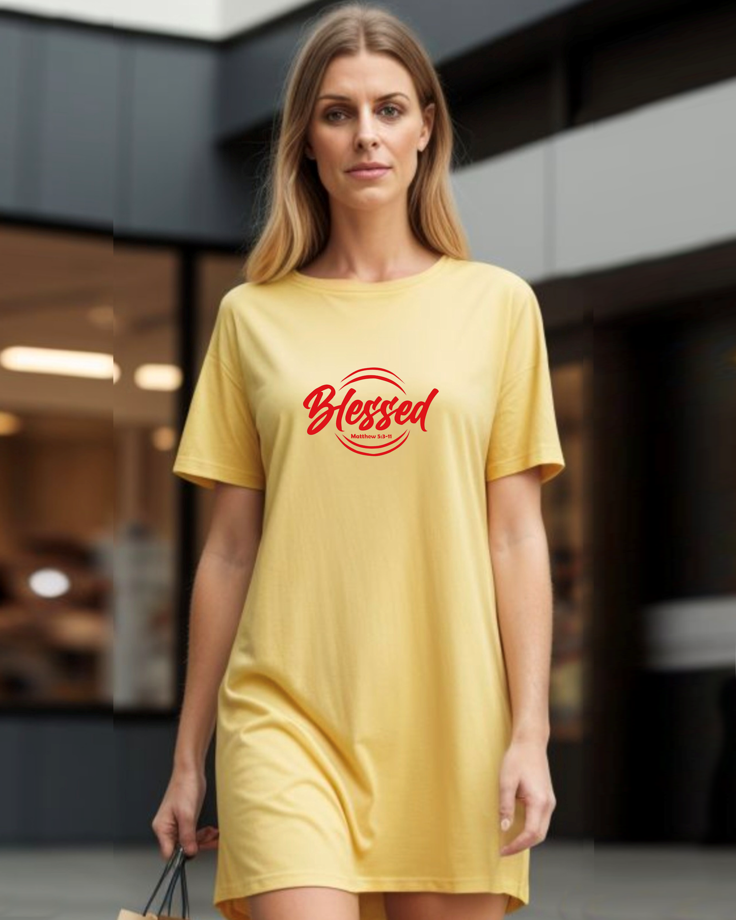 A woman in a shopping centre wearing a yellow Heavenly Dreamwear t-shirt dress with a red logo comprised of the text "Blessed" on top of a broken double circle with a biblical scripture reference beneath the text.