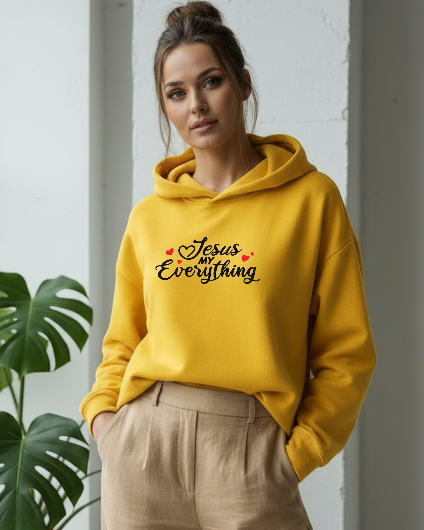A woman wearing a bright yellow hoodie with the text 'Jesus MY Everything' in black script and small red hearts, paired with beige trousers, standing indoors next to a green plant.