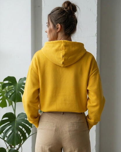 A woman wearing a bright yellow hoodie paired with beige trousers, viewed from the rear standing indoors next to a green plant.