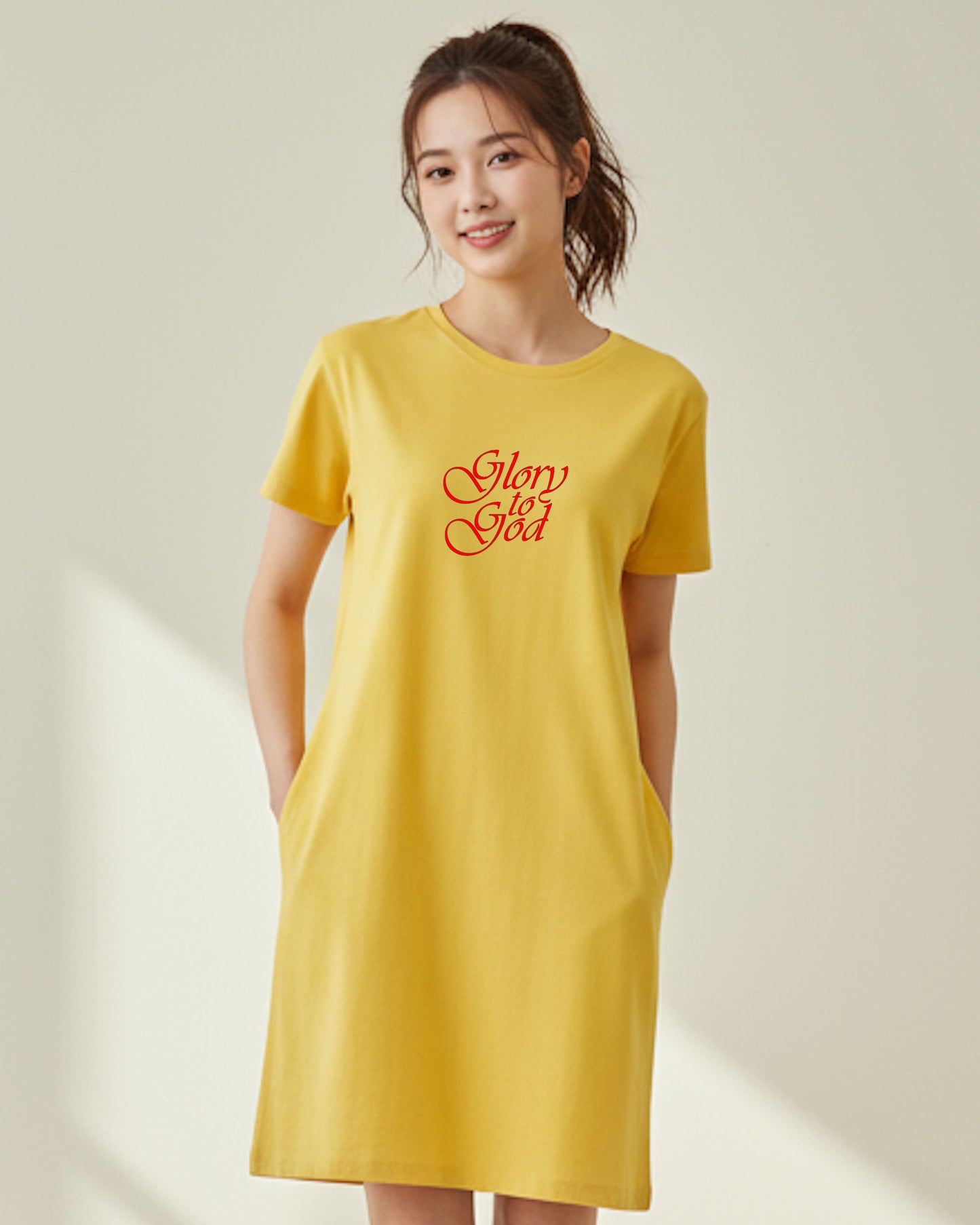 A woman indoors wearing a yellow t-shirt dress with red text the says, 'Glory to God' against a pale yellow wall