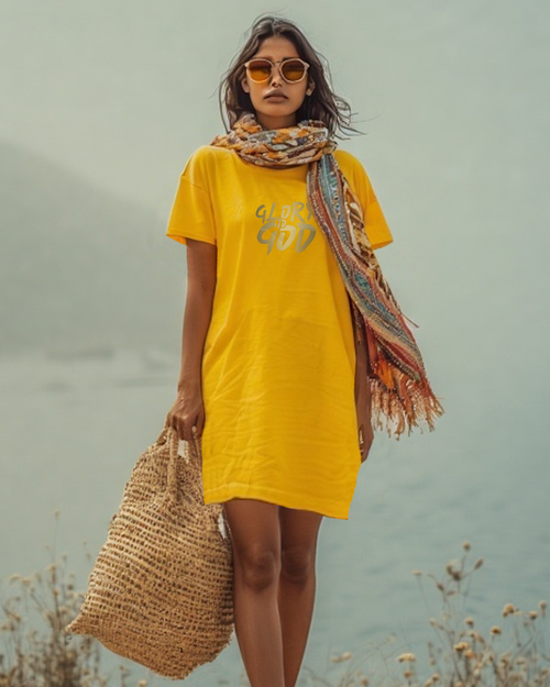 A woman on a hill wearing a scarf, a yellow Heavenly Dreamwear t-shirt dress with a gold "Gory to God" text logo printed on the front. She is carrying a large raffia bag and wearing a large scarf and sunglasses.