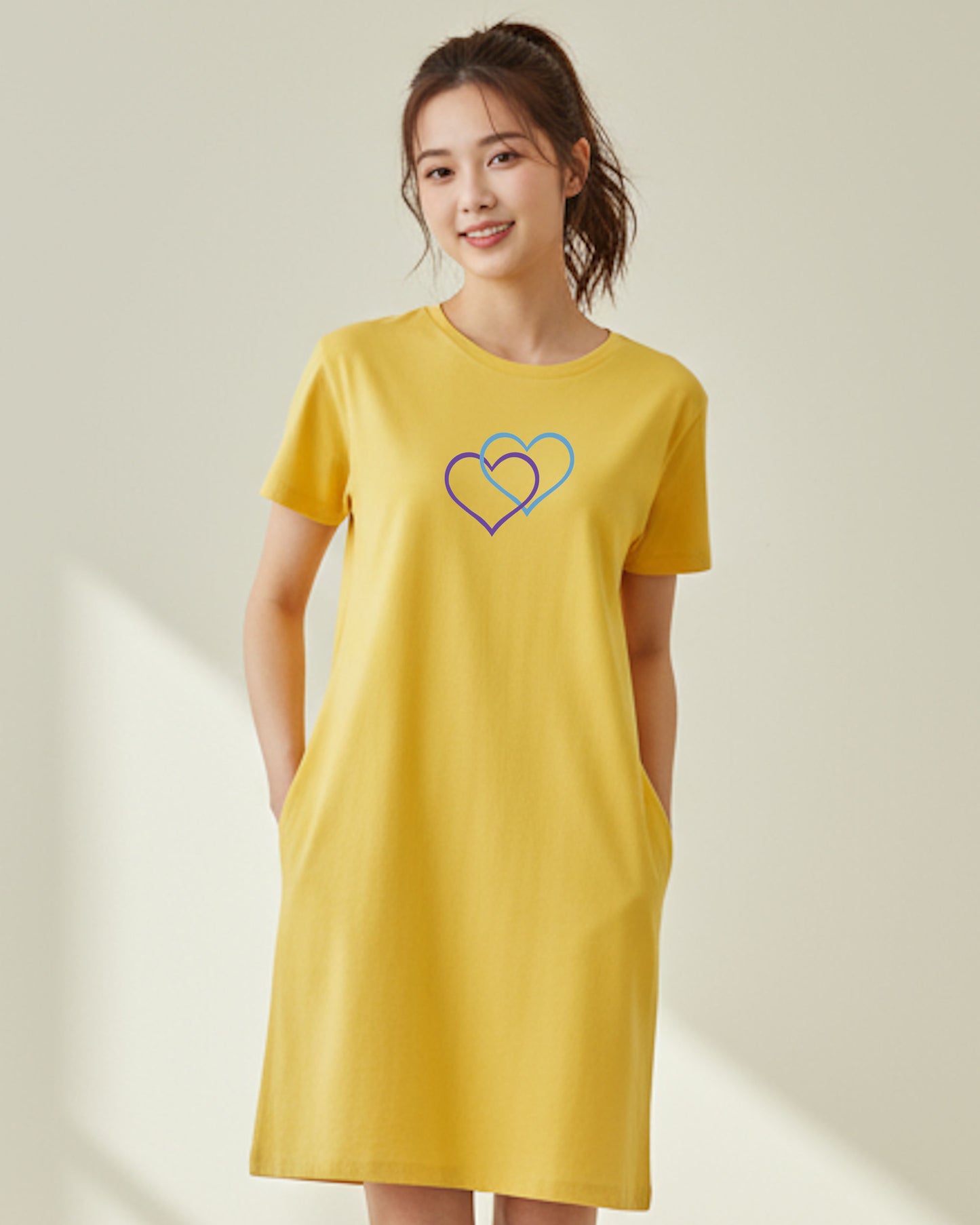 A woman wearing a yellow t-shirt dress with a Heavenly Dreamwear logo made of an purple and blue intertwined heart shapes on the front against a plain beige background.