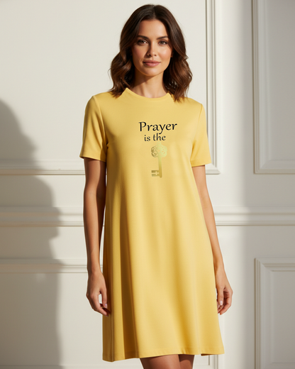 A woman wearing a yellow short-sleeve dress with the text 'Prayer is the key' and a gold key graphic on the front, standing against a white paneled wall.
