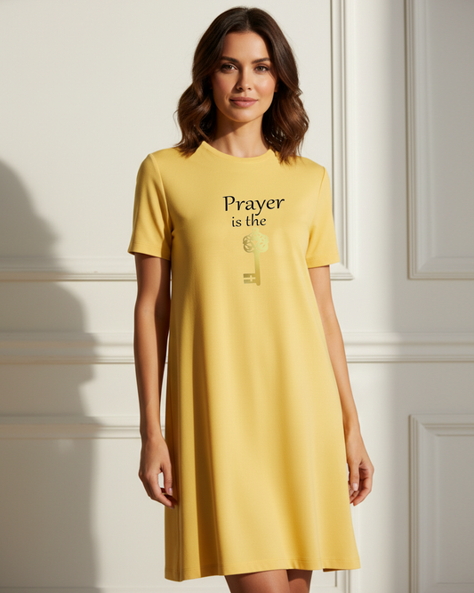 A woman wearing a yellow short-sleeve dress with the text 'Prayer is the key' and a gold key graphic on the front, standing against a white paneled wall.