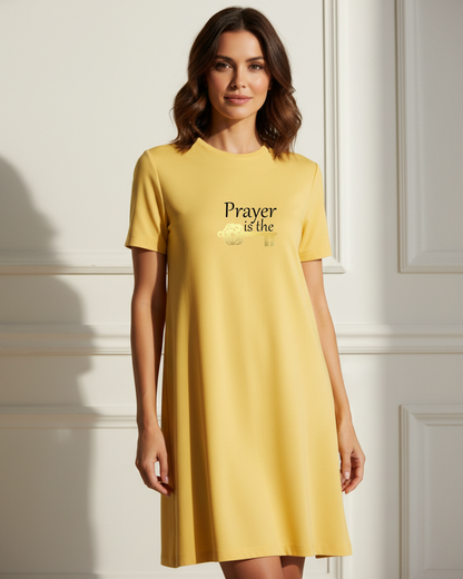 A woman wearing a yellow t-shirt dress featuring a logo of black text 'Prayer is the,' followed by a gold key symbol in a white wall panel room