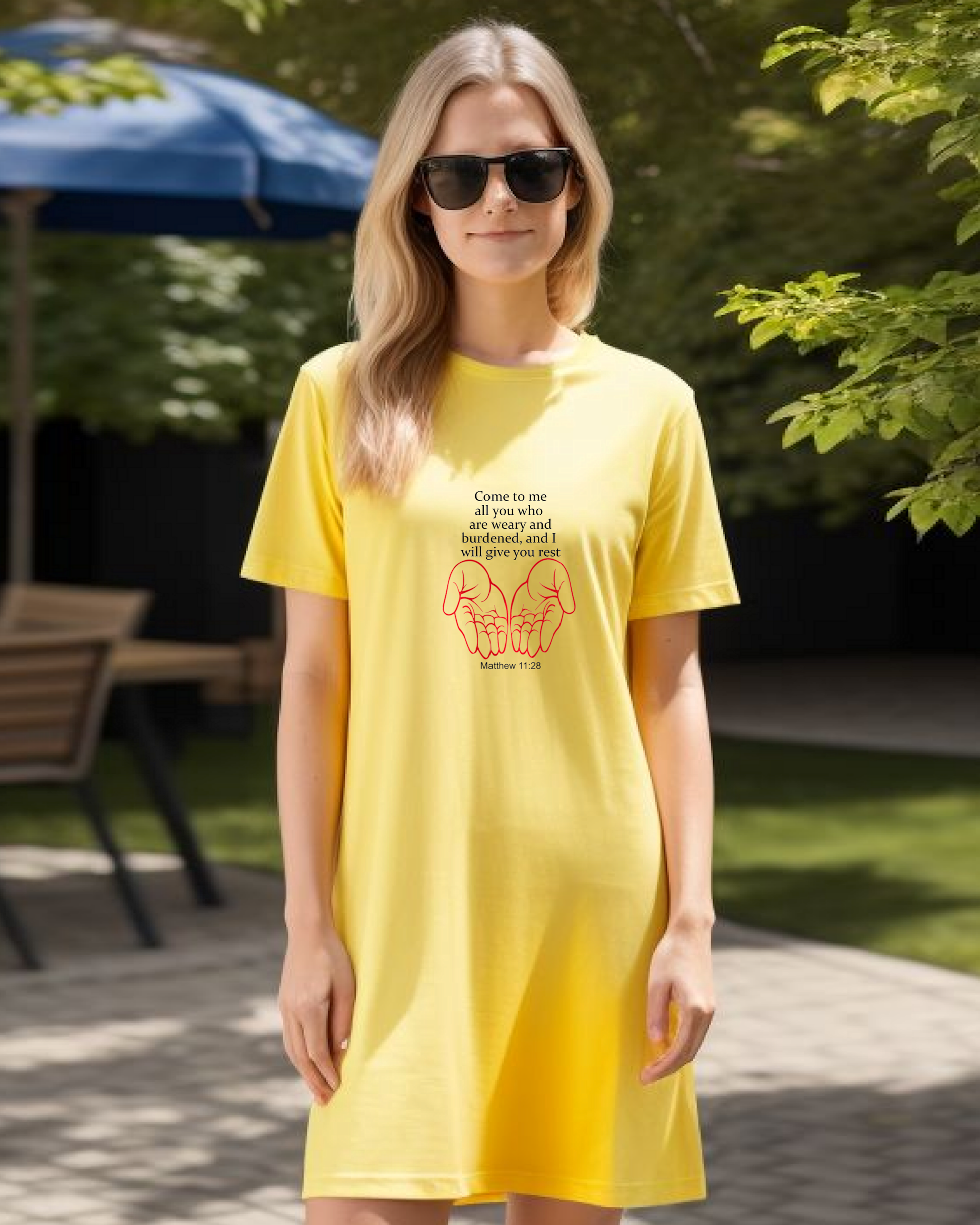 A woman in a garden wearing a yellow Heavenly Dreamwear t-shirt dress with black "come to me all you who are weary" text with two red hands facing palm up logo.