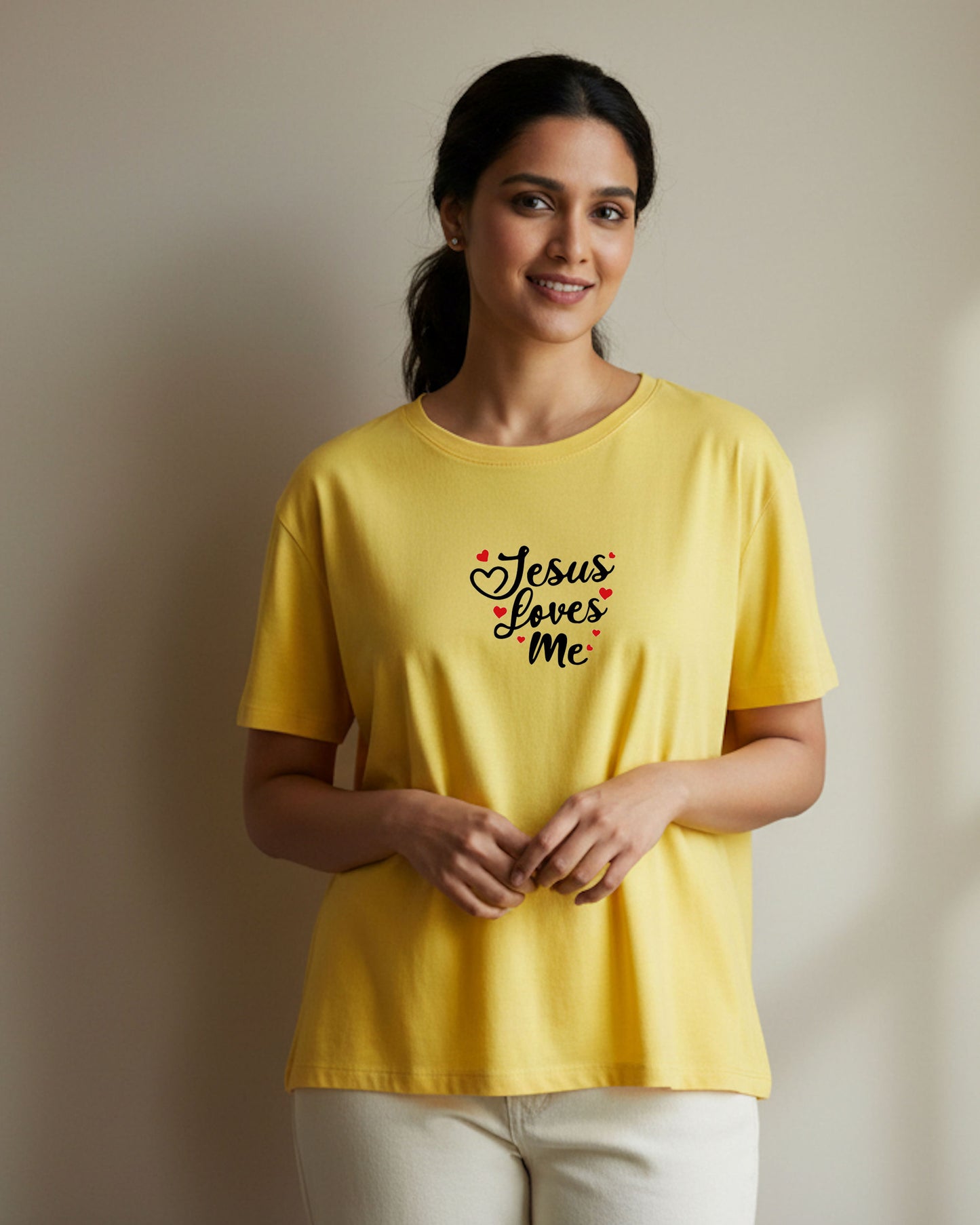 A woman wearing a yellow t-shirt with black 'Jesus Loves Me' text and red hearts against a plain yellow background
