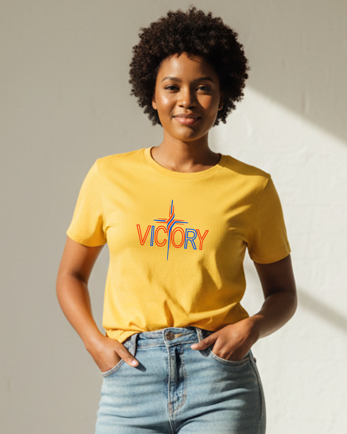 A woman with short dark curly hair wearing a yellow crewneck t-shirt with a colorful 'VICTORY' graphic on the chest, paired with light blue jeans, standing against a neutral light gray wall with warm sunlight casting a shadow.