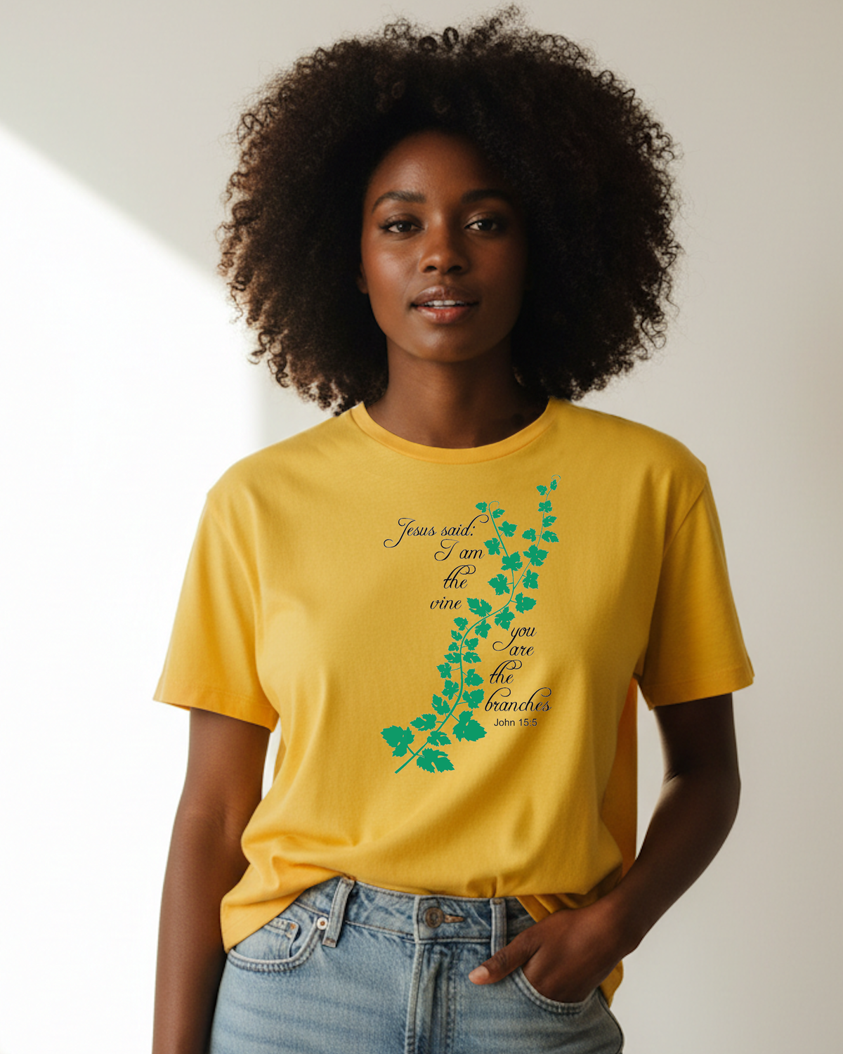 A woman with curly dark hair wearing a mustard yellow graphic t-shirt that has a green vine motif with black text 'Jesus said I am the vine you are the branches' printed, with light-wash blue jeans, standing indoors in a brightly lit room.