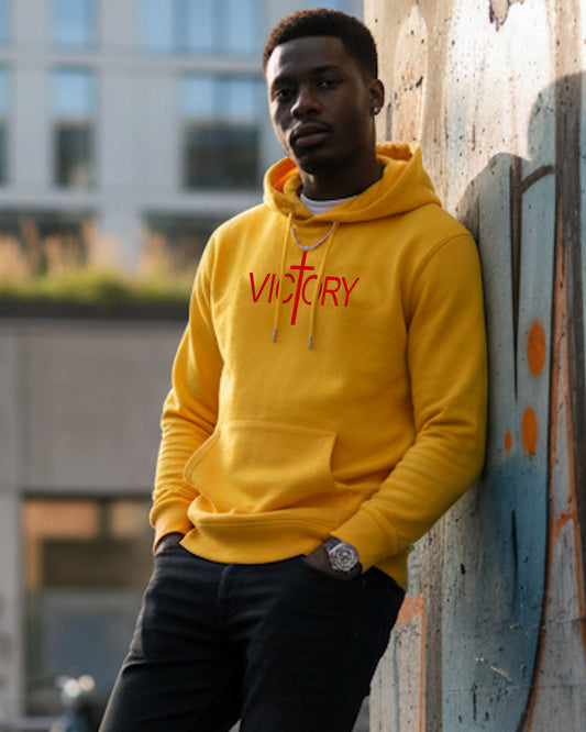 A young man wearing a yellow hoodie with red text 'VICTORY' printed on it, leaning against a wall.