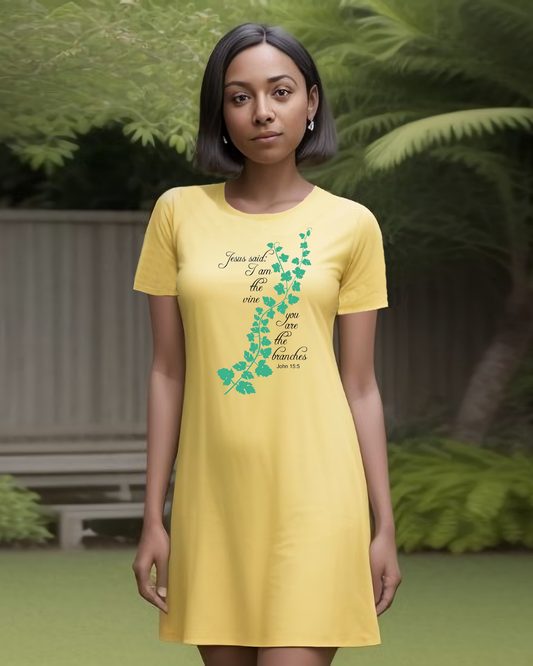 Woman standing in a garden wearing a yellow t-shirt dress with a logo made up of a green vine draped across the front with a biblical scripture on either side in black text from John 15:5