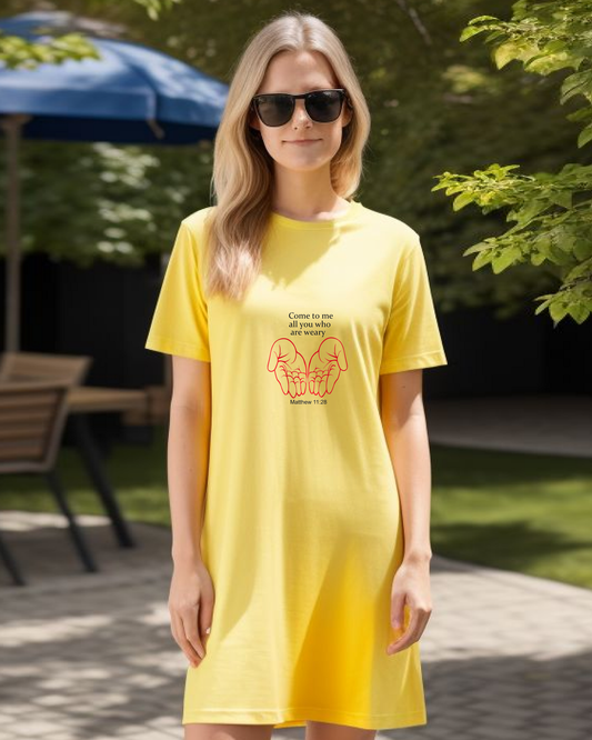 A woman standing in a garden wearing a yellow Heavenly Dreamwear t-shirt dress with a black "come to me all you who are weary" text with two red hands facing palm up logo printed on the front.