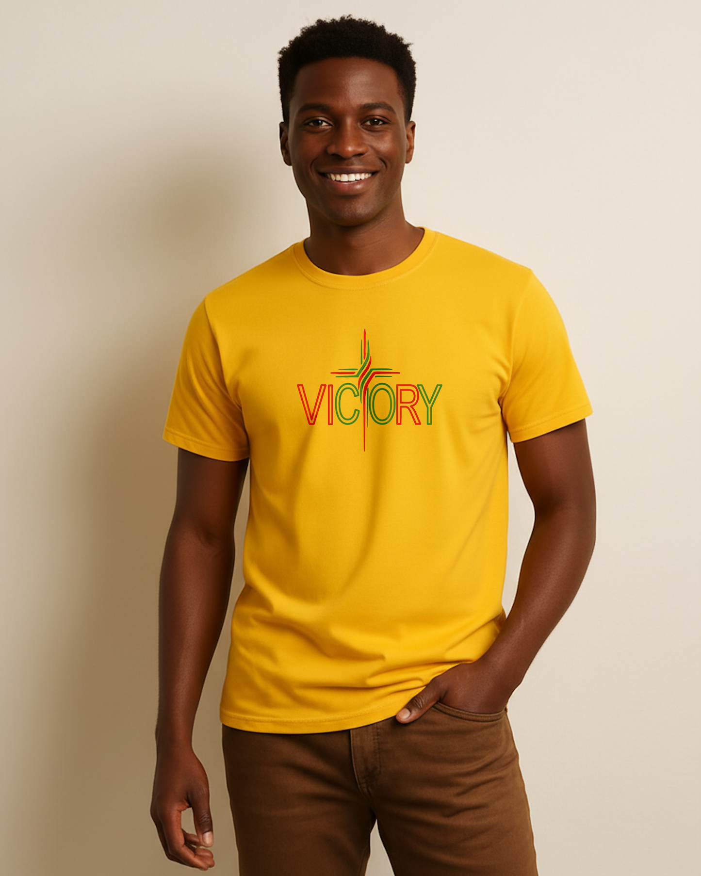A smiling man wearing a bright yellow t-shirt with a colorful red and green 'VICTORY' graphic on the chest, paired with brown pants, standing against a plain beige background.